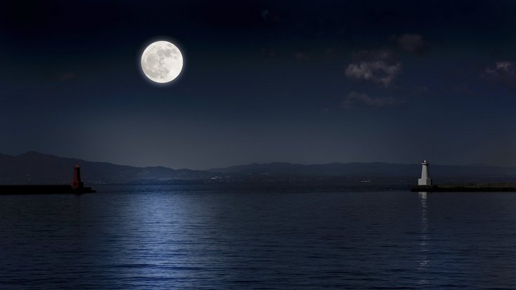 Foto einer Meeresbucht bei Nacht, links wirft der Vollmond weißes Licht auf die Wasseroberfläche, rechts steht ein heller Leuchtturm
