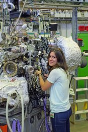 Eine junge Frau steht vor einem zirka zwei Meter hohen wissenschaftlichen Instrument. Sie blickt über ihre Schulter in Richtung Kamera, während sie an einer Schraube des Instruments hantiert. Am Instrument verlaufen viele Kabel, viele Instrumententeile davon sind mit silberner Folie abgedeckt.