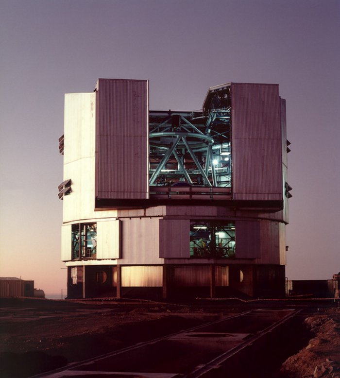 Eines der vier Teleskope des VLT Runde riesige glänzende Halle in der Wüste, Hallentor in der oberen Hälfte ist beidseitig zur Seite geschoben und geöffnet, ebenso das Dach. In der Halle wird ein Metallgerüst sichtbar: das Teleskop.