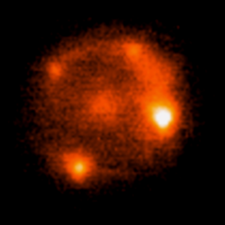 Vier unterschiedlich helle Objekte, ringförmig gruppiert um ein schwaches, ausgedehntes Objekt im Zentrum.