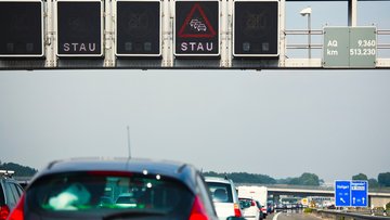 Unten im Bild stehen viele Autos auf einer Autobahn, vom Betrachter aus wegzeigend, hintereinander auf einer Autobahn. Oben ist eine schwarze Tafel, auf der „Stau“ steht und ein rotes Dreieck mit drei Autos abgebildet ist.