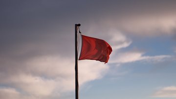 Eine rote Flagge weht im Wind an einer hohen Fahnenstange vor bewölktem Himmel mit vereinzelten blauen Lücken, im Gegenlicht der tiefstehenden Sonne. 