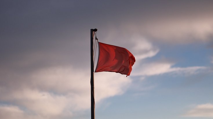Eine rote Flagge weht im Wind an einer hohen Fahnenstange vor bewölktem Himmel mit vereinzelten blauen Lücken, im Gegenlicht der tiefstehenden Sonne. 