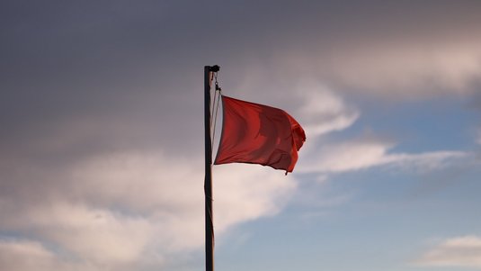 Eine rote Flagge weht im Wind an einer hohen Fahnenstange vor bewölktem Himmel mit vereinzelten blauen Lücken, im Gegenlicht der tiefstehenden Sonne. 