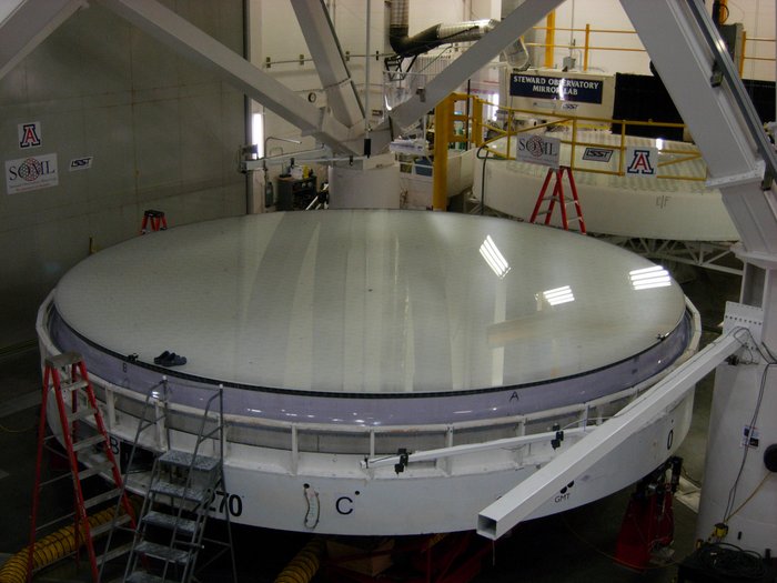 Der Blick in eine optische Fertigungshalle des Steward Observatory Mirrorlab zeigt eine
weißlich-glänzend schimmernde große Glasplatte. Sie liegt in einer Halterung, im Hintergrund ist die ursprüngliche Gussform zu
erkennen. Auf dem zukünftigen Spiegel des Giant Magellan Telescope stehen am Rand zwei Gummischlappen, die sich im Vergleich zum Spiegeldurchmesser winzig ausnehmen.