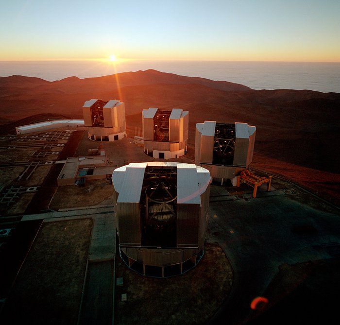 Luftbild eines Bergplateaus, auf dem vier kantige Gebäude zu sehen sind; durch große offene Tore sind die Gestänge der vier Teleskope zu sehen; im Hintergrund Berge der Atacama-Wüste mit tiefstehender Sonne.