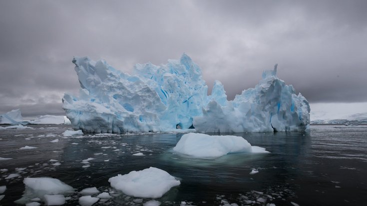 Eisberg in der Antarktis | Bildquelle: burroblando/iStock Das Foto zeigt einen großen, zerklüfteten Eisberg, der in dunklem Wasser schwimmt. Der Himmel ist bedeckt und grau, was eine kühle und etwas düstere Atmosphäre erzeugt.