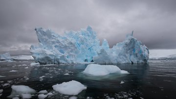 Das Foto zeigt einen großen, zerklüfteten Eisberg, der in dunklem Wasser schwimmt. Der Himmel ist bedeckt und grau, was eine kühle und etwas düstere Atmosphäre erzeugt.