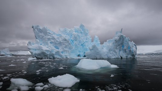 Das Foto zeigt einen großen, zerklüfteten Eisberg, der in dunklem Wasser schwimmt. Der Himmel ist bedeckt und grau, was eine kühle und etwas düstere Atmosphäre erzeugt.