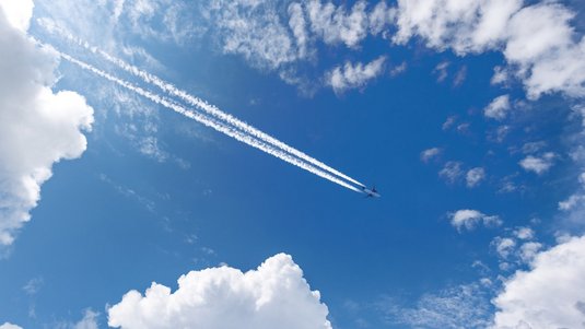 Blick von unten in strahlend blauen Himmel mit weißen Wolkenfeldern am Rand: Ein Flugzeug fliegt diagonal von oben links nach unten rechts und zieht zwei parallele, lange weiße Kondensstreifen hinter sich her.