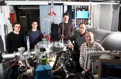 Gruppenfoto der Forscher um zylindrische, silberne Vakuumkammer.