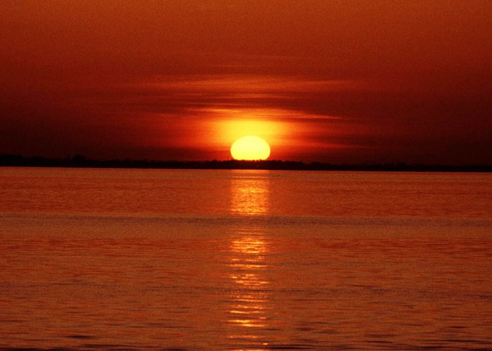 Nur noch ein kleiner Teil der Sonnenscheibe ragt über dem Horizont. Am Himmel sind angestrahlte Wolkenstreifen zu erkennen.