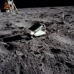 Fotografie, Spiegel auf der Mondoberfläche, durch die Halterung ist er schräg nach oben gerichtet. Auf der Spiegelfläche sind die einzelnen Katzenaugen als kleine Kreise zu erkennen. Im Hintergrund ist das Raumschiff der Astronauten zu sehen.