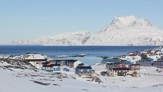 Eine Ortschaft im Schnee an einer Küste, im Hintergrund große, felsige Berge