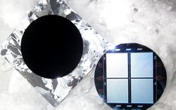 Links liegt die Vorderseite der Solarzelle aus schwarzem Silizium als ein dunkler Kreis inmitten eines Rechtecks, das silbrig glänzt. Daneben ist die Rückseite, ein Kreis mit vier Rechtecken, auf denen jeweils senkrechte Linien sind.