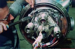Foto eines Mannes, der an einer metallischen Röhre arbeitet. Diese enthält vier kleinere Röhren und zahlreiche verkabelte technische Komponenten.