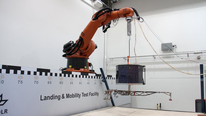 Eine mit Solarzellen bestückte tonnenartige Konstruktion mit einem Gitter-Untergestell hängt an einem Roboterarm.