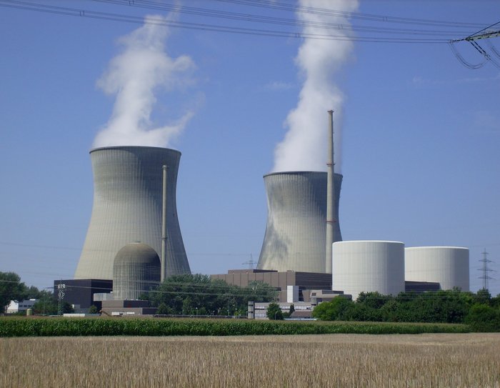 Dominant ragen die zylindrischen Kühltürme über ein Rapsfeld auf. Aus ihnen steigt Wasserdampf. Kleiner sind im Vordergrund die ebenfalls zylindrischen Betonbauten der Sicherheitsbehälter zu sehen.