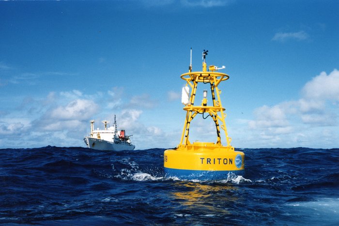 Foto. Offenes Meer. Eine Boje, im Wasser dümpelnd. Auf der Boje sind Messinstrumente zu erkennen. Ein Schiff im Hintergrund.