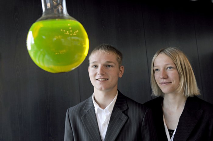 Zwei Schüler stehen vor einem Reagenzglas mit grünlich leuchtendem Inhalt.