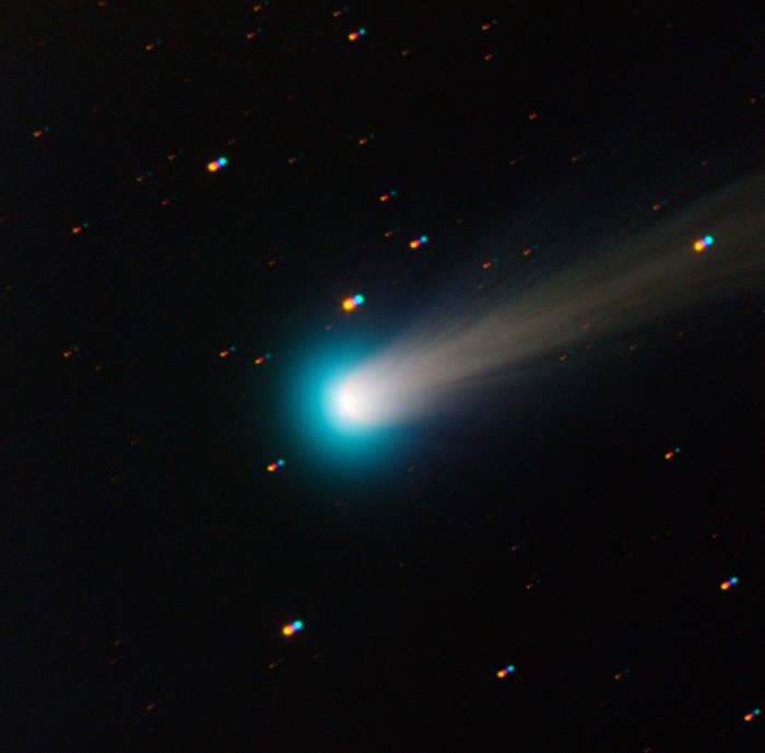 In der Mitte des Bildes ein hell leuchtender Fleck (der Komet), gefolgt von einem schwach leuchtenden Schweif, auf dunklem Hintergrund. Im Hintergrund sind außerdem einige kleine leuchtende Punkte zu sehen (Sterne).