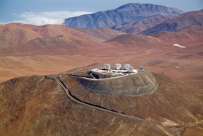 Der Paranal Blick von oben auf eine karge Landschaft, auf einer Bergerhebung Gebäude, im Hintergrund weitere Berge.