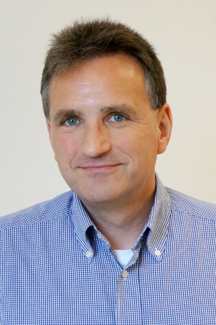 Alfons Khoukaz Porträt eines lächelnden Mannes in einem karierten Hemd.