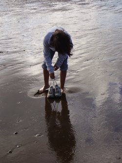Dieses Foto zeigt eine Frau, die barfuss im feuchten Sand steht. Rund um ihre Füße wirkt der Sand heller und er reflektiert das Licht nicht so gut wie der Rest des Sandes. Der Sand ist also trockener.