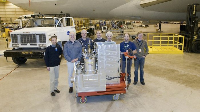 GREAT-Empfänger vor dem Transport ins Flugzeug Sieben Männer stehen hinter dem GREAT-Empfänger. Dieser ist ein etwa 1,50m hoher metallener Kasten mit einem zylinderförmige Aufbau.