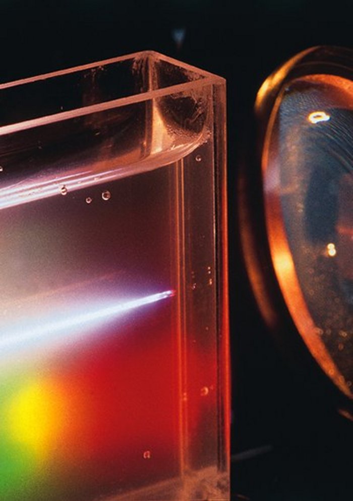 Laser steuert chemische Reaktion Foto: rechts Linse, links schmaler mit einer klaren Flüssigkeit gefüllter Behälter, in dem Behälter ein heller Strahl, der sich von rechts nach links etwas auffächert.
