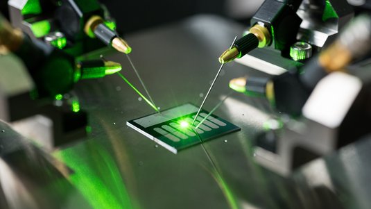 Auf einer Glasplatte liegt ein Mikrochip und wird mit Laserlicht bestrahlt.