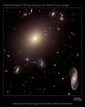 Blick in einen Galaxienhaufen. Viele kleine Strudel aus Licht, wie Nebelfetzen. Davor hunderte von Sternen. Im linken oberen Bereich ein großes, elliptisches, nach außen hin schwächer werdendes Lichtwölkchen.