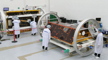 Foto eines Laborraums, in dem ein Satellit in eine Halterung eingespannt ist. Personen in Schutzkleidung untersuchen den Satelliten. 