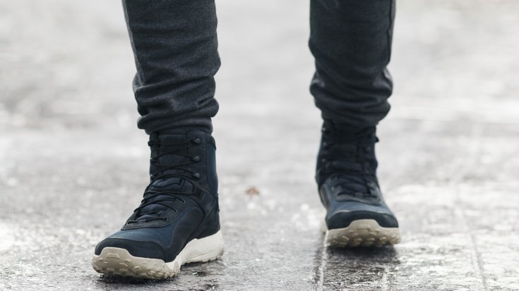 Person mit dunklen Winterstiefeln steht auf einer vereisten, rutschigen Fläche. Die Szene wirkt kalt und winterlich, nur die Beine und Schuhe sind sichtbar.