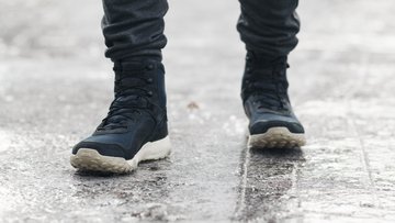 Person mit dunklen Winterstiefeln steht auf einer vereisten, rutschigen Fläche. Die Szene wirkt kalt und winterlich, nur die Beine und Schuhe sind sichtbar.