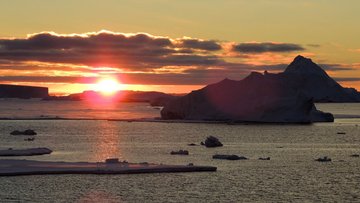 Meereslandschaft am Südpol, im Vordergrund einige Felsen und Eisschollen im Meer, im Hintergrund geht die Sonne unter