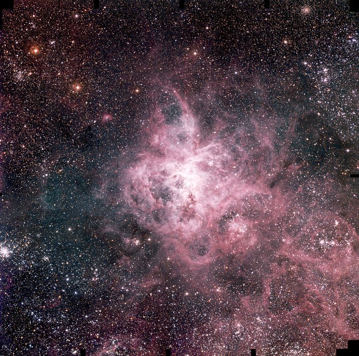Tarantel-Nebel Himmelsaufnahme übersät von Sternen; in der Mitte helle zartrosa Gasmassen, die an die Form einer Spinne erinnern.