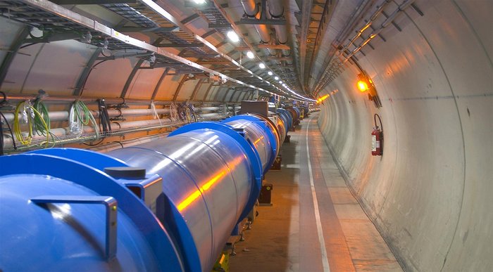 Blick in den LHC-Tunnel Foto: Blick entlang einer leicht nach rechts gekrümmten Betontunnelröhre, in der ein Teilchenbeschleuniger aus langen, metallenen Röhren mit zahlreichen Kabeln und anderen technischen Geräten aufgebaut ist.