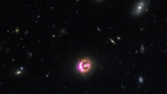 Viele Galaxien, die Galaxie in der Bildmitte ist von einem Ring umgeben, in dem sich vier helle Strahlungsquellen befinden.