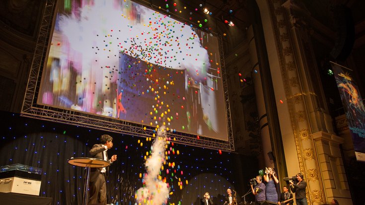 Highlights-Show mit Ranga Yogeshwar, Bildquelle: Highlights der Physik/Uhmeyer; Lizenz: gemäß den Bedingungen der Quelle Ranga Yogeshwar betrachtet eine Explosion von bunten Kunststoffbällen auf der Highlights-Bühne.