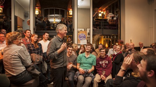 Ein Wissenschaftler hält einen Vortrag in einer Bar, während er von Menschen umgeben ist.