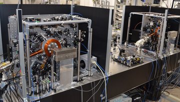 Auf einem Tisch im Labor stehen zwei senkrechte Platten mit vielen Spiegeln und Optiken, die die Atomuhren sind.