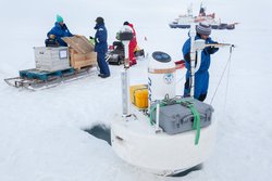 Foto. Auf einer verschneiten Eisscholle öffnen vermummte Menschen zwei Holzkisten, die sich auf einem Schlitten befinden. Im Vordergrund steht eine Boje neben einem Loch im Eis. Auf der Boje sind verschiedene Sensoren installiert. In der Ferne ist ein Forschungsschiff zu erkennen.