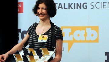 Eine junge Frau steht vor einem Bühnenhintergrund, auf dem in verschiedenen Größen das FameLab-Logo zu sehen ist. Sie hält ein großes Modell mit drehbaren Spiegeln in der Hand und lächelt.