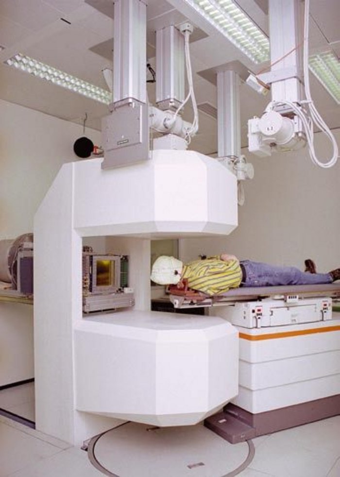 PET-Kamera integriert in die Bestrahlungsanlage Weißer Behandlungsraum, zentral circa drei Meter hoher Rahmen, an dem oben und unten zwei einen Meter dicke abgerundete weiße Blöcke befestigt sind, wie Ober- und Unterteil eines Brötchens. Rechts in mittlerer Höhe liegt Patient mit Gipsmaske auf Bahre, Oberkörper und Kopf ragen genau in Freiraum zwischen den Blöcken hinein. Links davon glänzende Metallfläche, Kabel und weitere Technik. Von der Decke ragen senkrechte Träger mit Kabel und Gerätschaften bis in drei Meter herunter.