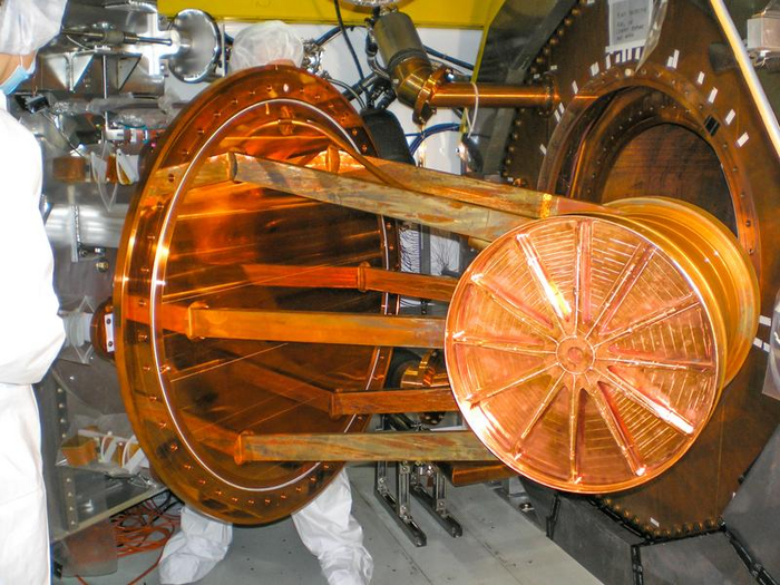 EXO-200 Detektor Rechts im Bild die runde Öffnung zu einer Kammer, davor der schwere große runde Deckel, beides aus kupferfarbenem Metal. Links im Bild ein Mensch in Schutzkleidung aus weißem, weitem Stoff.