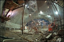 Baumaßnahmen in einer gigantisch großen Halle mit Betonwänden. Die Bauarbeiter wirken winzig, die Gerüste reichen mehrere Stockwerke hoch. 