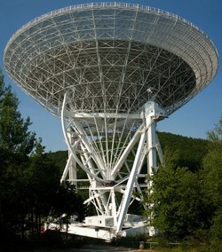 Großes, weißes Teleskop, dessen Schüssel gen Himmel gerichtet ist. Es befindet sich freistehend auf einer Waldlichtung.