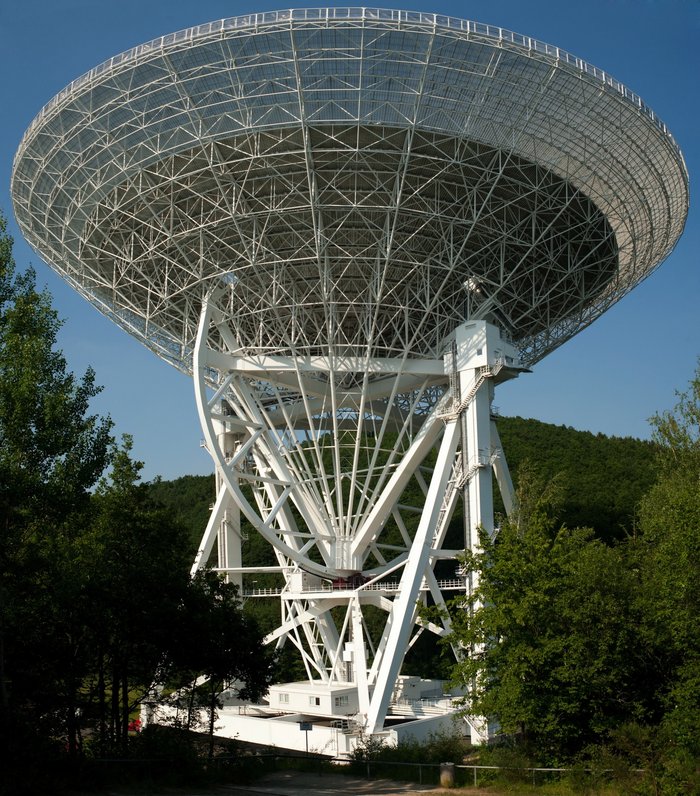 Großes, weißes Teleskop, dessen Schüssel gen Himmel gerichtet ist. Es befindet sich freistehend auf einer Waldlichtung.