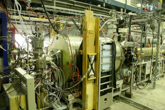 Experimentierhalle des ALPHA Experiments Ein sehr komplizierter Aufbau aus technischen Geräten in einer Halle. Im Zentrum ist ein großer messingfarbener Zylinder zu sehen, darum herum viele Drähte und Kabel.